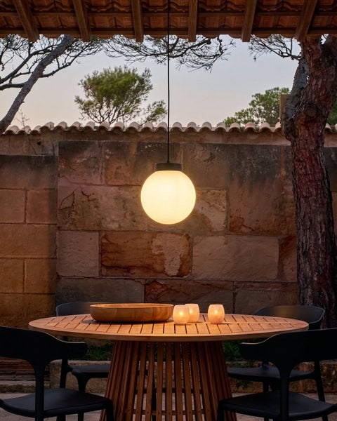 Eine moderne kugelförmige Pendelleuchte hängt über einem runden Holztisch mit Stühlen auf einer Terrasse vor einer Steinmauer.