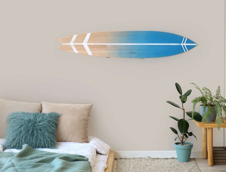 Wanddekoration aus Holz in Surfbrett-Form über einem Bett mit Kissen und Decke neben Zimmerpflanzen in einem hellen, modern eingerichteten Raum.