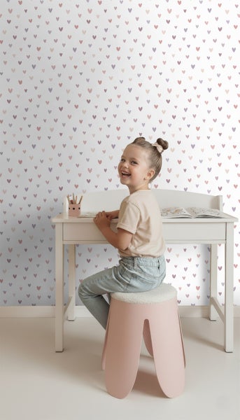 Mädchen sitzt an weißem Schreibtisch vor einer Wand mit Herzchenmuster