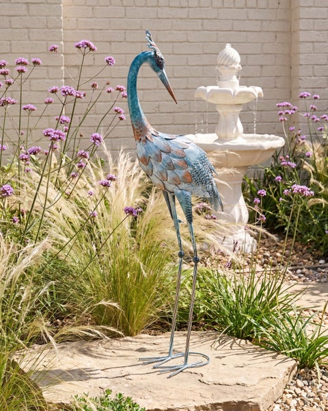 Hell gestalteter Garten mit einer Gartenfigur aus Metall in Form eines Reihers, Ziergräsern, Blumen und einem Etagenbrunnen.