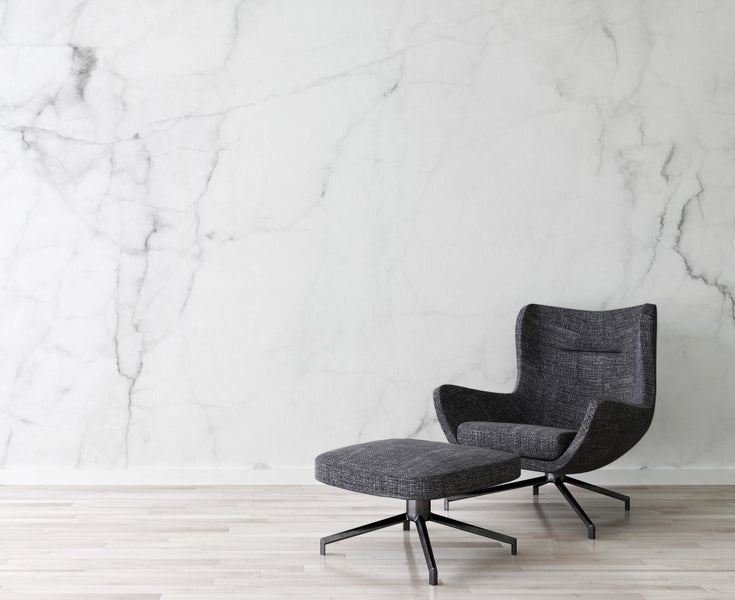 Moderner grauer Sessel mit Hocker vor einer Wand in Marmoroptik und hellem Holzboden.