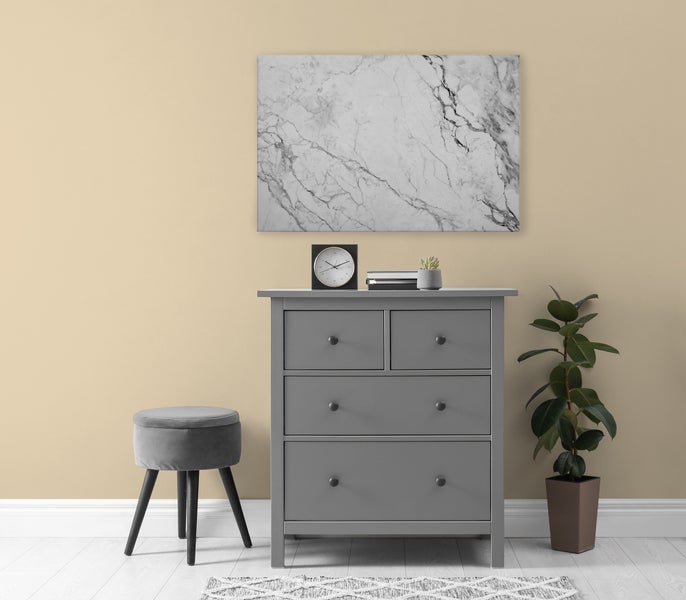 Eine graue Kommode mit drei Schubladen steht vor einer beigen Wand mit einem Marmor Wandbild, daneben ein Hocker und eine Zimmerpflanze.