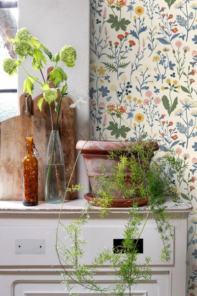 Vliestapete mit buntem Wildblumen-Muster im Landhausstil, dekoriert auf weißem Sideboard mit Pflanzen, Glasflaschen und Holz-Schneidebrettern.