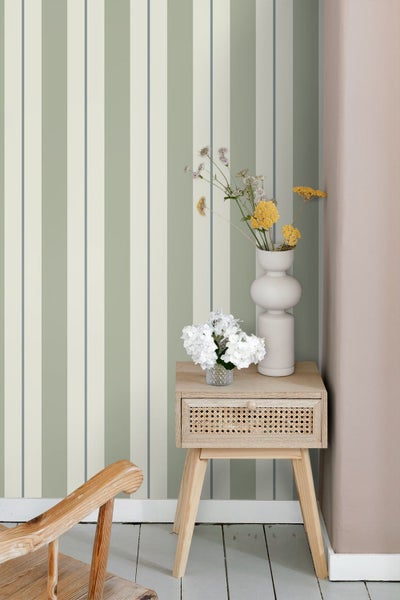 Gestreifte Tapete in Salbeigrün und Creme hinter einem Nachttisch aus Holz mit Rattanschublade und Blumenvasen in einem hellen Zimmer.