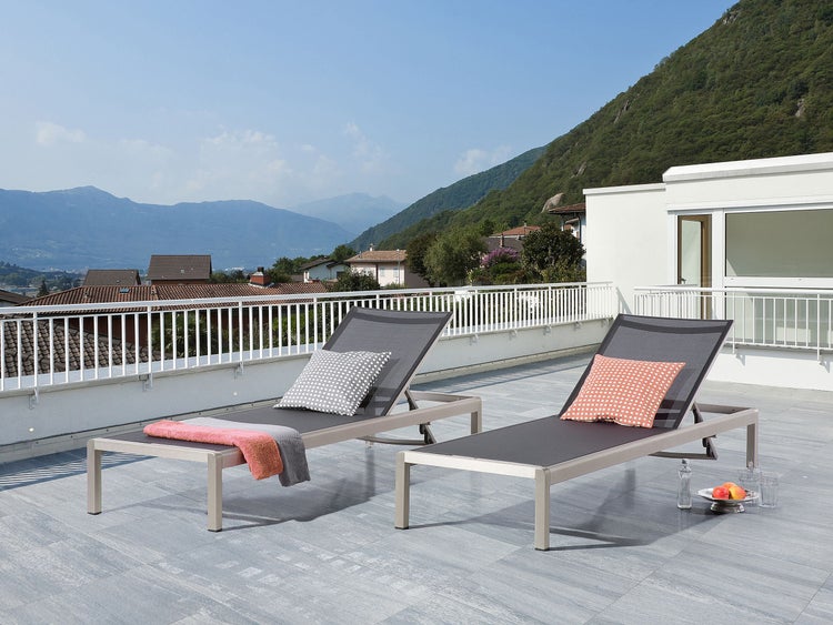 Zwei graue Sonnenliegen mit Kissen und Getränken auf einer Terrasse mit Blick auf die Berge.