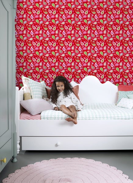 Mädchen sitzt auf einem Kinderbett vor einer Wand mit gemusterter Tapete in einem Kinderzimmer.