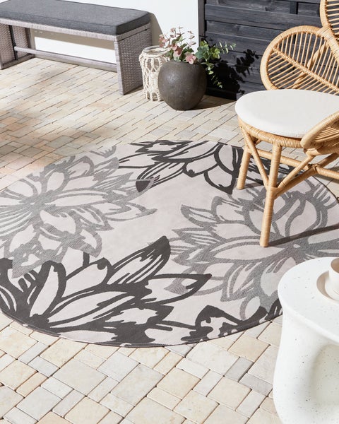Runder Outdoor-Teppich mit Blumenmuster auf einer Terrasse aus Stein mit Rattan-Stuhl und Sitzbank in einer hellen, organisierten Umgebung.