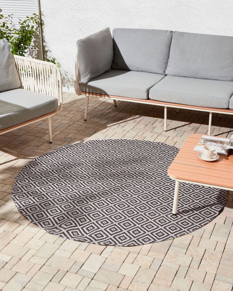 Moderner Außenbereich mit rundem Outdoorteppich mit Rautenmuster, grauem Loungesofa, Sessel und Holztisch auf einer hellen Steinterrasse.