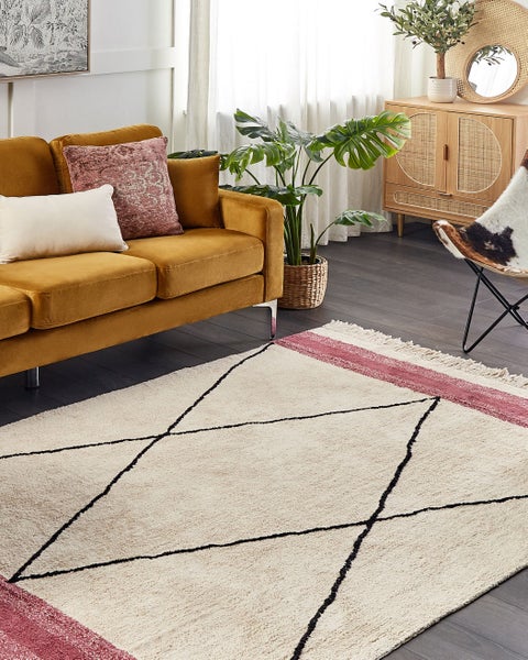 Hochflor-Teppich mit schwarzem Rautenmuster und rosa Streifen in einem Wohnzimmer mit gelbem Samtsofa, Monstera-Pflanze und Holzkommode.