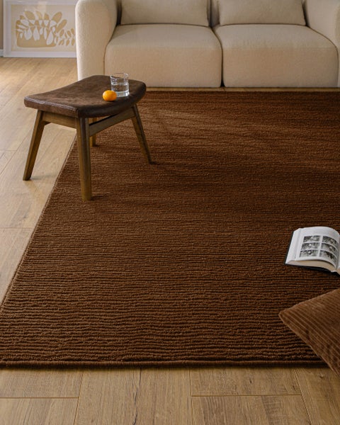 Gerippter brauner Teppich in hellem Wohnzimmer mit Sofa, Holzfußboden, einem Hocker aus Holz und Leder sowie einem Kissen.