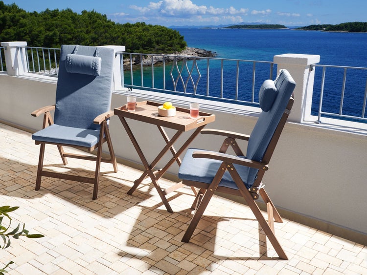 Gartenmöbel-Set aus Holz auf einer hellen Terrasse mit Meerblick, bestehend aus zwei Klappsesseln mit Auflagen und einem klappbaren Tisch.