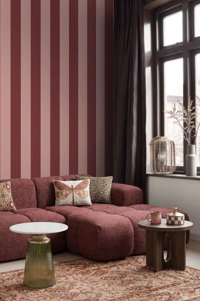 Wohnzimmer mit rot-rosa gestreifter Tapete, burgunderfarbenem Modulsofa, Beistelltischen und gemustertem Teppich.