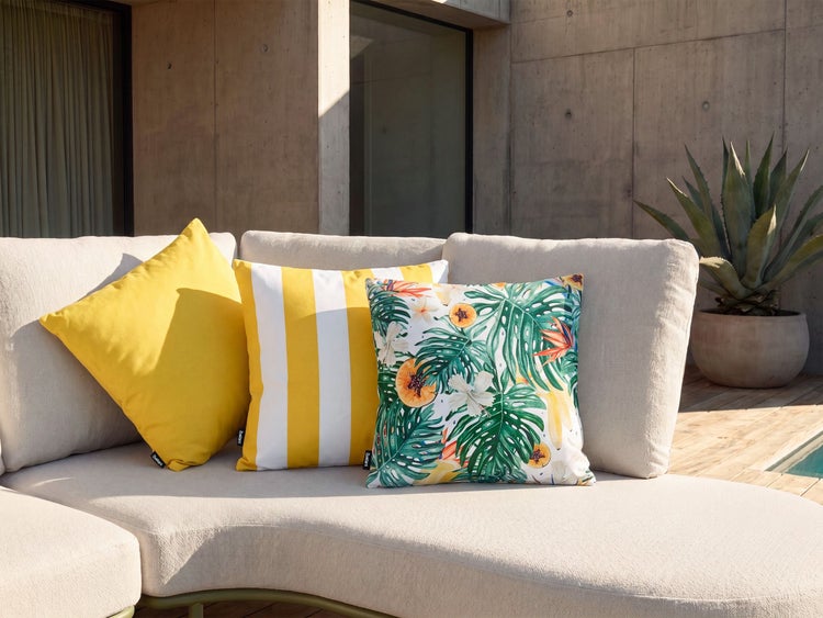 Drei dekorative Gartenkissen in verschiedenen Mustern auf einem hellen Lounge Sofa vor einer modernen Betonwand mit einer Agavenpflanze.