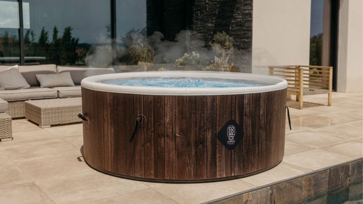 Runder aufblasbarer Whirlpool in Holzoptik mit dampfendem Wasser auf einer modernen Terrasse mit Gartenmöbeln. COCO SPA Logo.