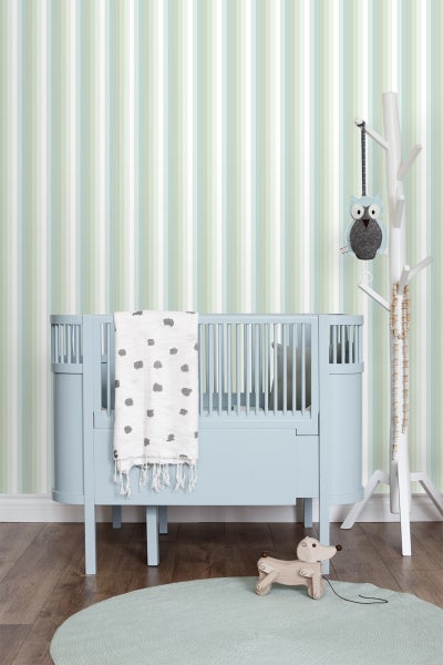 Ein Kinderzimmer mit einem blauen Gitterbett, einer Decke mit Punktmuster, einem Spielzeug-Hund und einer Wand mit Streifenmuster.