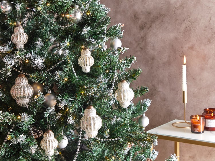Weihnachtsbaum geschmückt mit gerippten Glasanhängern, silbernen Kugeln, Perlengirlande und warmweißer Lichterkette vor einer strukturierten Wand.