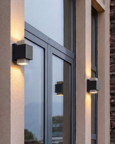 Moderne Außenwandleuchten in Würfelform mit Up- und Downlight an einer Hausfassade neben großen Glasfenstern.