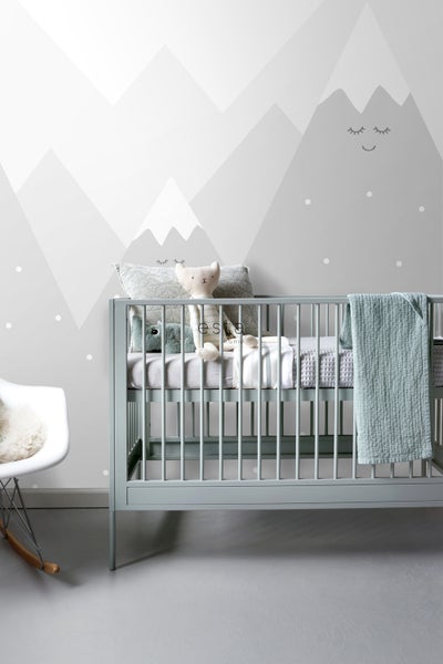 Babyzimmer mit Gitterbett, Kissen und Stofftieren vor einer Wandtapete mit Bergmotiv.