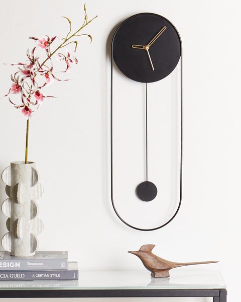 Moderne Wanduhr mit Pendel und Metallrahmen in einem hellen, minimalistischen Zimmer mit Vase, Orchidee und Holzfigur.