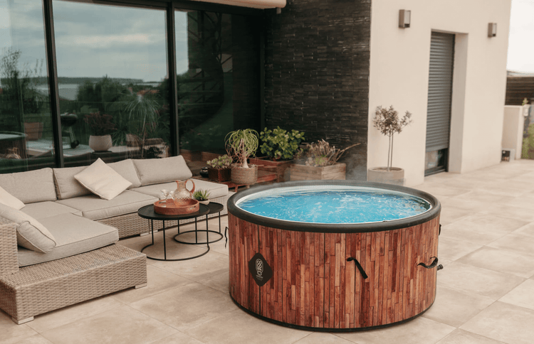 Runder Whirlpool mit Holzoptik und Coco Spa Logo auf einer modernen Terrasse mit Loungemöbeln und Pflanzen.