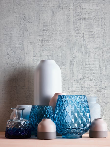 Dekorative Szene mit Zimmerpflanze, Hausmodellen und gerahmtem Bild auf einem Beistelltisch vor einer gemusterten Wand.