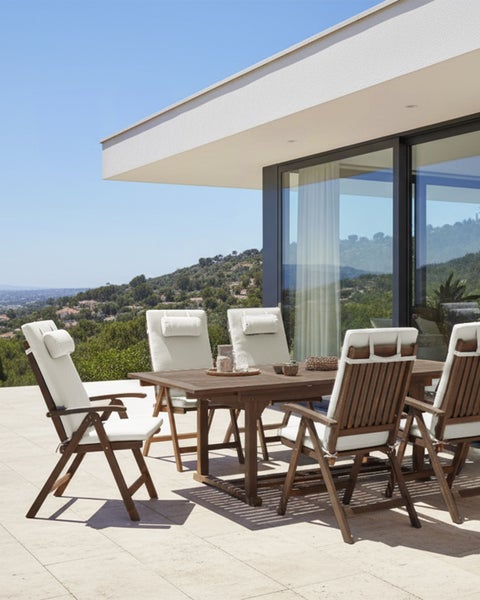 Helles Gartenmöbel-Set aus Holz mit Tisch und Stühlen inklusive weißen Auflagen auf einer organisierten Terrasse mit Ausblick.