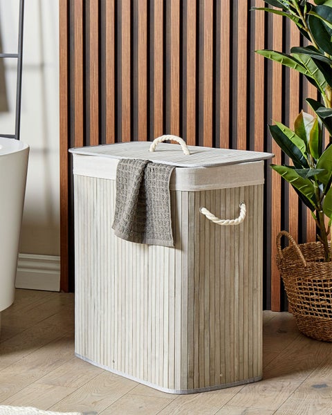 Wäschekorb aus hellgrauen Holzlamellen mit Kordelgriffen und Deckel in einem modernen Badezimmer mit Holzpaneelwand.