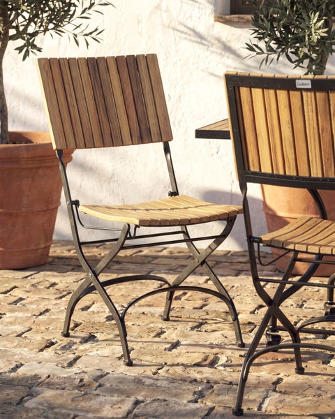 Klappbarer Gartenstuhl aus Holzlatten und schwarzem Metallgestell auf einer gepflasterten Terrasse neben einer weißen Wand.