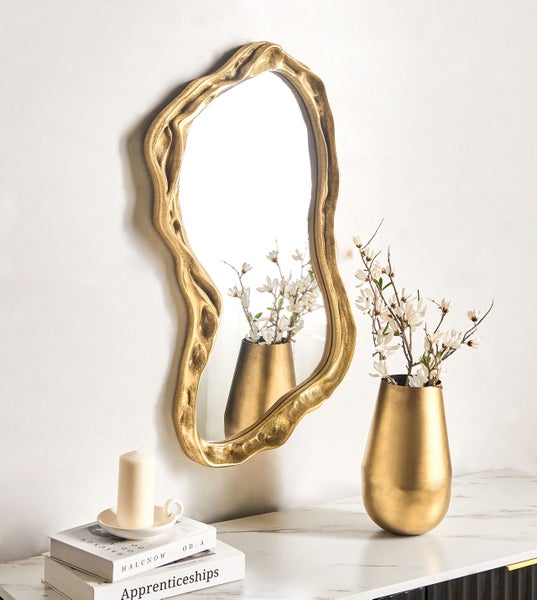 Wandspiegel in organischer, unregelmäßiger Form mit goldfarbenem Metallrahmen auf einer hellen Marmoroberfläche mit Vase und Kerze.