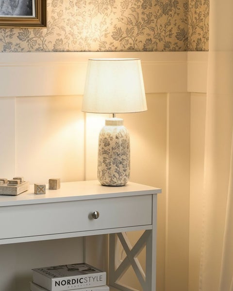 Klassische Tischleuchte mit Keramikfuß mit Blumenmuster und Stoffschirm auf einer Kommode vor einer gemusterten Tapete in einem hellen Zimmer.