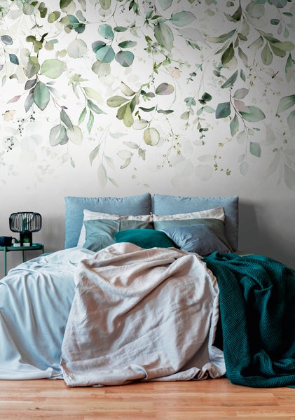 Botanische Fototapete mit Eukalyptusblättern in einem hellen Schlafzimmer mit Holzboden, weichen Textilien und grünen Kissen.