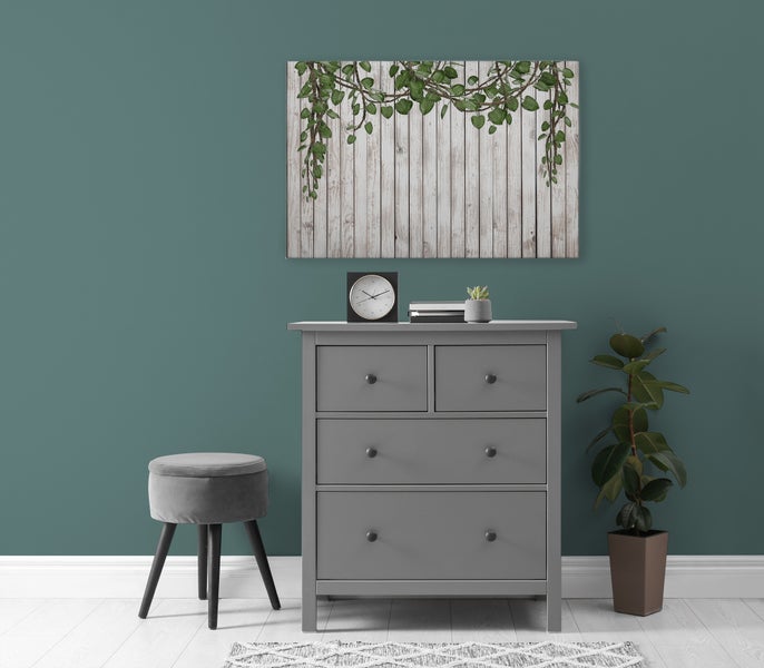 Wandbild mit Motiv einer Holzwand mit hängenden Pflanzen über einer grauen Kommode mit Hocker und Pflanze in einem modern eingerichteten Raum.