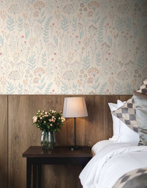 Schlafzimmer mit blumiger Vliestapete und einem Bett mit hölzernem Kopfteil, Nachttischlampe und Blumenstrauß in der Vase.