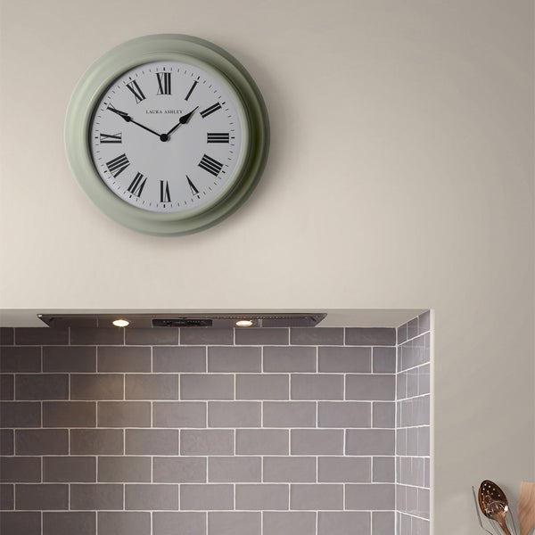 Runde Laura Ashley Wanduhr mit römischen Ziffern in einer hellen Küche über einem Fliesenspiegel und einer Dunstabzugshaube.