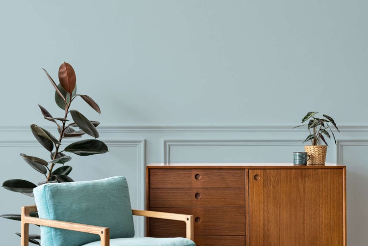 Hellblaue Wandfarbe in einem hellen Wohnzimmer mit Wandleisten, Sideboard aus Holz, blauem Sessel und Zimmerpflanzen.