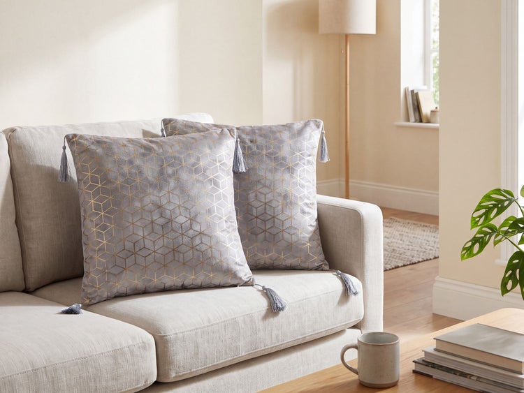 Zwei quadratische Dekokissen mit geometrischem Muster und Quasten auf einem Sofa in einem hellen Wohnzimmer mit Stehlampe und Pflanze.