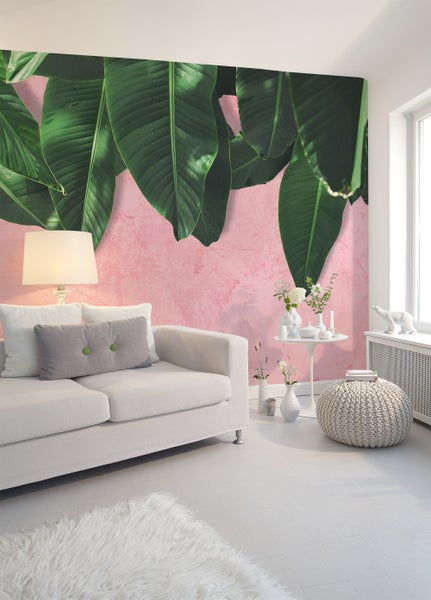 Fototapete mit großen grünen Bananenblättern auf rosa Hintergrund in einem hellen Wohnzimmer mit weißem Sofa und Strickhocker.
