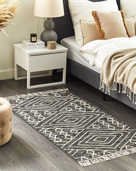 Schlafzimmer mit geometrisch gemustertem Läufer vor einem Bett und weißem Nachttisch auf dunklem Holzboden.