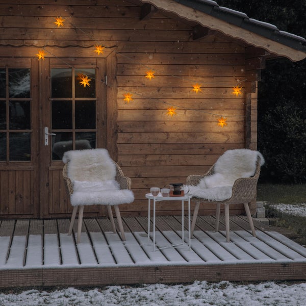 Winterliche Szene mit Holzhütte, Lichterkette in Sternform, zwei Stühlen mit Fellauflage und einem kleinen Tisch mit Tee