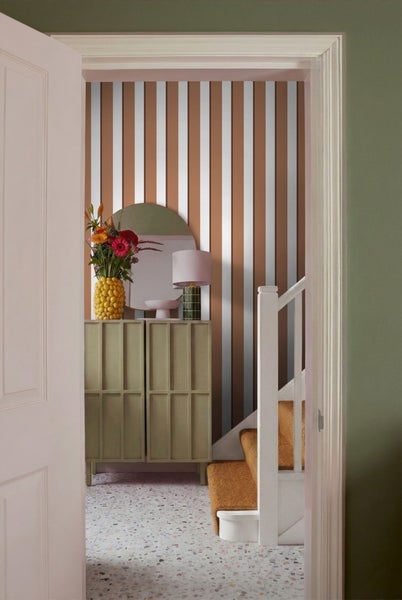 Blick durch eine offene Tür auf einen Flur mit gestreifter Tapete, Kommode, Spiegel, Blumenvase, Treppe und Teppich