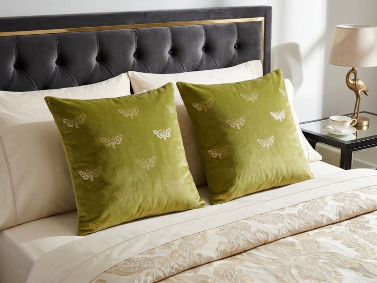 Zwei grüne Zierkissen aus Samt mit eingestickten Schmetterlingsmotiven auf einem hellen Bett vor einem dunkelgrauen, gepolsterten Kopfteil.