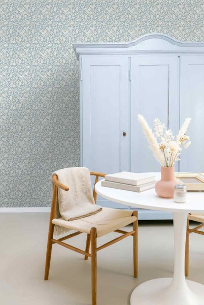 Vliestapete mit Blumenmuster in einem hellen Zimmer mit blauem Schrank, weißem Rundtisch und einem Holzstuhl mit Decke.