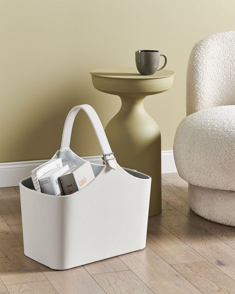 Weißer Aufbewahrungskorb aus Kunstleder mit Büchern in hellem Wohnzimmer mit modernem Beistelltisch, Tasse und Bouclé-Sessel auf Holzboden.