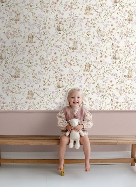 Vliestapete im Kinderzimmer mit Hasen und Blumenmuster, ein Kind sitzt auf einer Holzbank vor der tapezierten Wand.