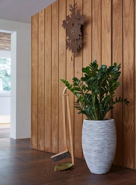 Zimmerpflanze in weißem Pflanzkübel, Kehrgarnitur aus Holz und Kuckucksuhr vor einer Wand mit vertikalen Holzlamellen.
