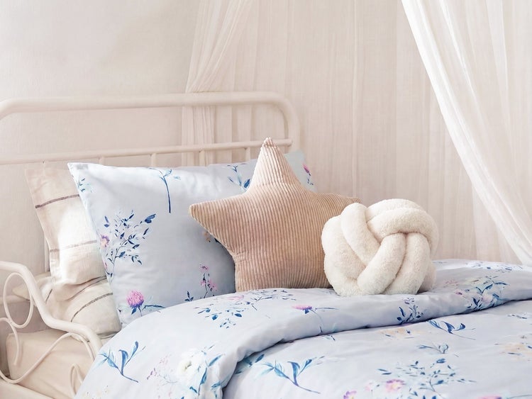 Ein helles Schlafzimmer mit Metallbett Bettwäsche mit Blumenmuster sowie einem sternförmigen und einem knotenförmigen Zierkissen.