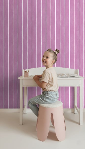 Mädchen sitzt an einem Schreibtisch vor einer gestreiften Tapete in einem Kinderzimmer.