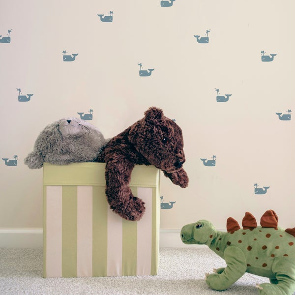 Kinderzimmer mit Wandtattoos in Form von Walen, einer gestreiften Aufbewahrungsbox mit Stofftieren und einem Plüschdinosaurier auf dem Teppich.