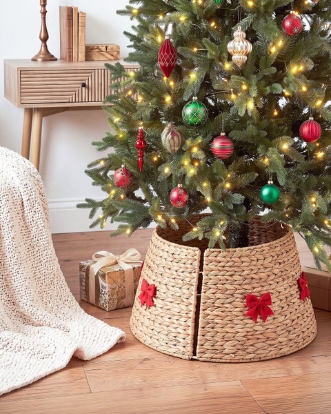 Weihnachtsbaum mit Lichtern und Kugeln in einem geflochtenen Baumständer aus Wasserhyazinthe mit dekorativen Schleifen im Wohnzimmer.
