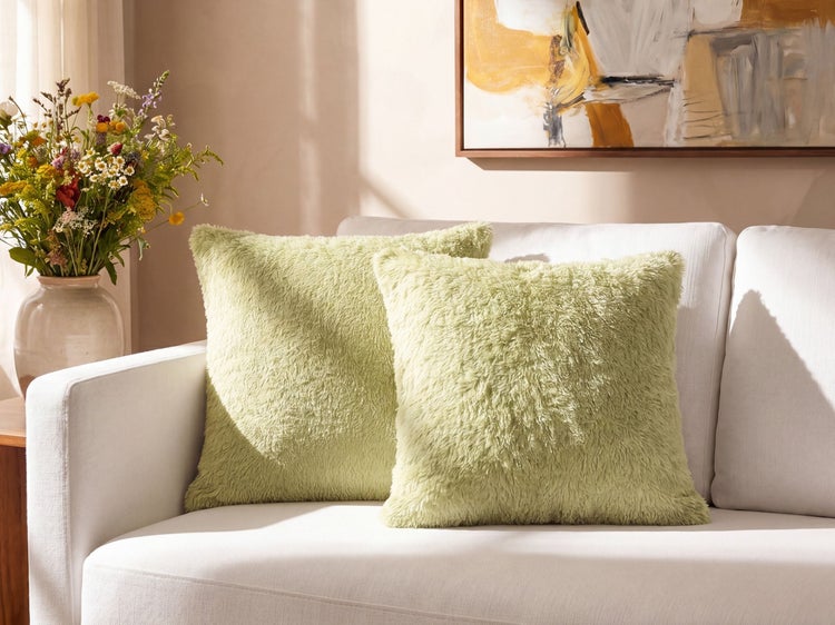 Zwei quadratische, flauschige grüne Dekokissen auf einem Sofa in einem hellen Wohnzimmer neben einer Blumenvase und einem Wandbild.